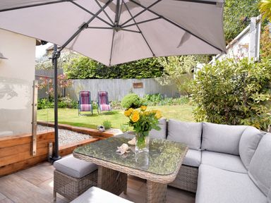 A patio with a glass table and wicker seating under a large umbrella overlooking a garden with two striped chairs at Bucket & Spade Lyme Regis