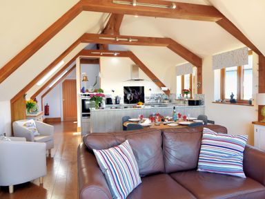 An open plan living space with a brown leather sofa and striped cushions a dining table set for six and a kitchen with wooden beams and windows at Bucket & Spade in Lyme Regis