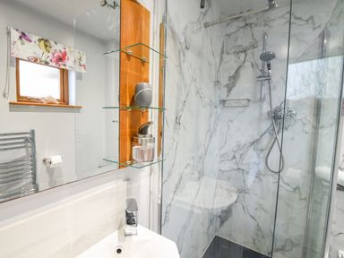 A bathroom with a corner sink, glass shelves, a window with floral curtains, a towel rack, and a marble-patterned glass shower enclosure at Bucket & Spade Lyme Regis
