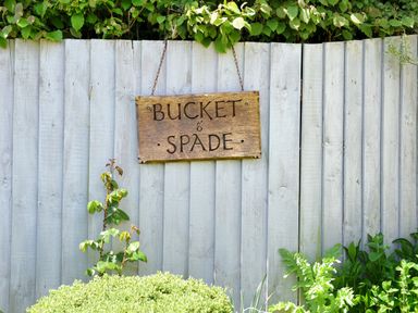 A wooden sign reading bucket and spade hanging on a light grey wooden fence with green plants in front at Bucket & Spade in Lyme Regis