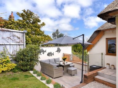 An outdoor patio area with a wicker sofa set and umbrella near a thatched roof house at Bucket & Spade in Lyme Regis
