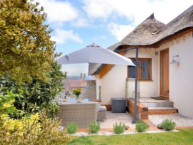An outdoor patio with wicker furniture and a large umbrella next to a thatched roof house at Bucket & Spade in Lyme Regis