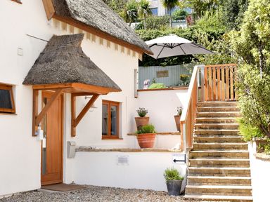 An exterior view of a building with a wooden door under a thatched roof canopy and stone steps leading to a terrace with an umbrella and potted plants at Bucket  Spade Lyme Regis