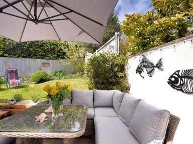 An outdoor seating area with a glass table umbrella and gray cushions next to a garden and white wall with fish decorations at Bucket & Spade in Lyme Regis