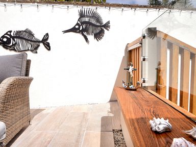 An outdoor seating area with a wicker chair a wooden bench with shells and an anchor decoration and metal fish wall art at Bucket & Spade Lyme Regis
