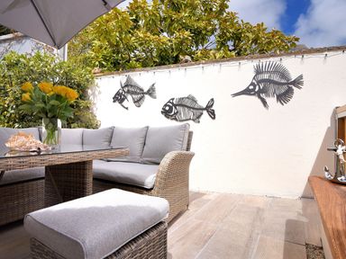 An outdoor seating area with a wicker sofa and table with flowers and seashell on the table and metal fish sculptures on a white wall at Bucket & Spade in Lyme Regis
