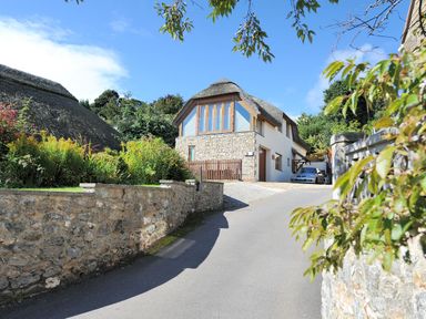 A house with stone walls and a thatched roof beside a driveway with a car behind a wooden fence at Bucket & Spade Lyme Regis