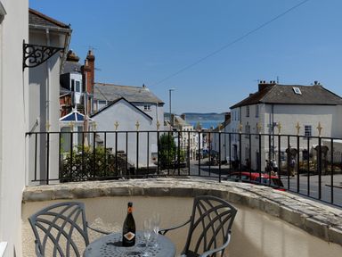 A balcony with a table and chairs overlooking a street with buildings and a view of the sea at Blacksmiths Cottage in Lyme Regis