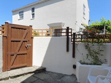A white building with a brown wooden gate and a small garden area with plants and a white plastic chair at Blacksmiths Cottage in Lyme Regis