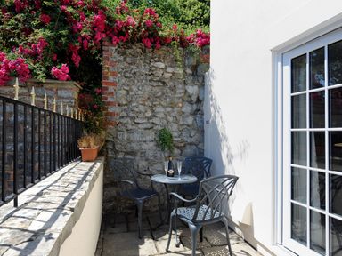An outdoor patio area with a small round table and three metal chairs next to a stone wall with red flowers at Blacksmiths Cottage in Lyme Regis