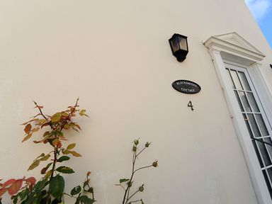A white wall with plants a black lamp a black sign reading Blacksmiths Cottage and a door at Blacksmiths Cottage in Lyme Regis