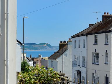 A coastal street with white buildings and a view of the sea and hills in the background at Blacksmiths Cottage in Lyme Regis