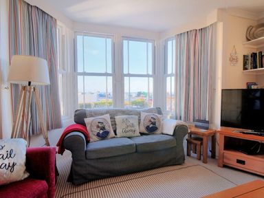 A living room with a blue sofa and patterned cushions by bay windows with striped curtains at 1 Cobb View in Lyme Regis