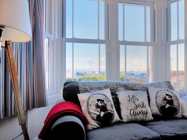 A living room with a grey sofa covered with three cushions and a red blanket near large windows with striped curtains at 1 Cobb View in Lyme Regis