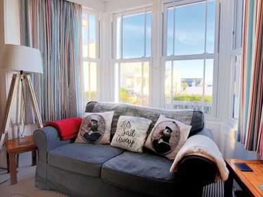 A sofa with decorative cushions and throws beside large windows with striped curtains and a floor lamp at 1 Cobb View Lyme Regis