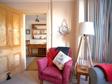 A living room with a red armchair and a wooden floor lamp next to a window with striped curtains at 1 Cobb View in Lyme Regis