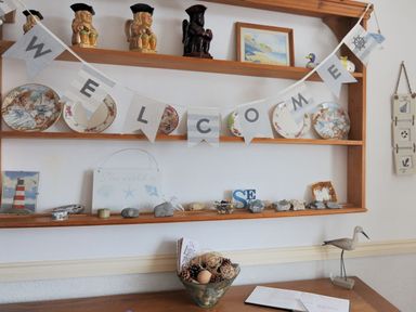 Wooden shelves with decorative plates figurines and welcome sign above a table with a bowl and an open notebook at 1 Cobb View in Lyme Regis