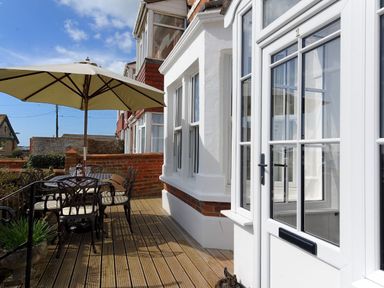An outdoor patio with a table and chairs under a large umbrella next to a white house with windows and a glass door at 1 Cobb View in Lyme Regis