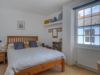A bedroom with a wooden bed frame a white bedspread pillows a teddy bear a window a radiator bedside tables lamps bookshelves and framed pictures at Riverview in Lyme Regis