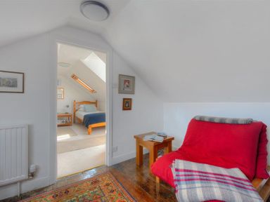 A small sitting area with a red covered chair and wooden side table next to a doorway leading to a bedroom with a wooden bed at Riverview in Lyme Regis