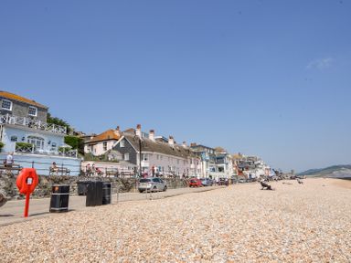 A pebble beach with people sitting on chairs and a row of houses along a promenade at 3 Argyle House in Lyme Regis