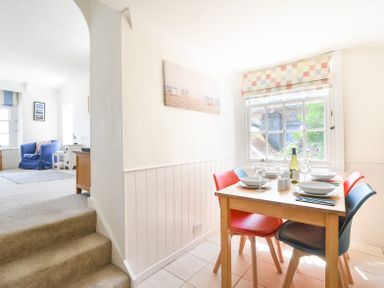 A dining area with a wooden table set with dishes and wine glasses near a window and a living room with a blue armchair and carpet at 3 Argyle House Lyme Regis