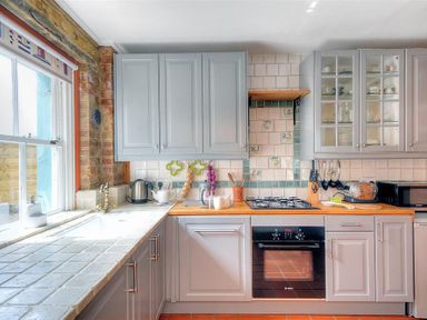 A kitchen with grey cabinets a window with a patterned blind a sink kettle and oven at Bedrock in Lyme Regis