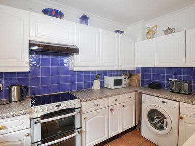 A kitchen with white cabinets blue tiled backsplash an electric stove microwave toaster washing machine kettle and knives at 2 Kersbrook Gardens in Lyme Regis