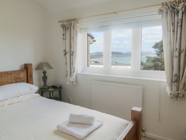 A bedroom with a wooden bed frame towels on the bed a side table with a lamp and a window with curtains overlooking a sea view at 2 Kersbrook Gardens Lyme Regis