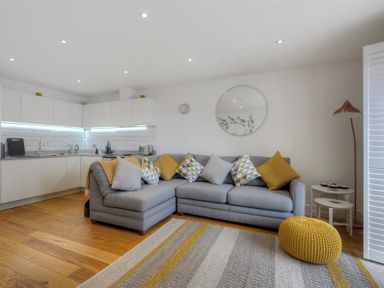 A living room with a grey sectional sofa with patterned and yellow cushions a knitted yellow pouf a patterned rug and a white kitchen with cabinets and appliances at The Lower Deck in Lyme Regis