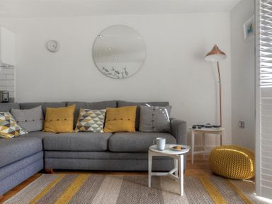 A living room with a gray sofa yellow and patterned cushions a round mirror a floor lamp and a small white table with a cup and plate at The Lower Deck in Lyme Regis