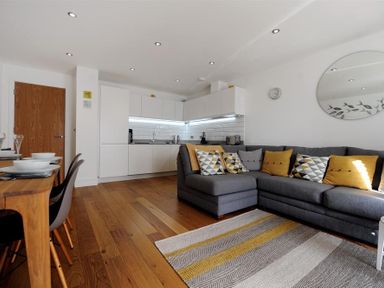 A kitchen with white cabinets and a dining table next to a grey sofa with yellow and patterned cushions at The Lower Deck in Lyme Regis