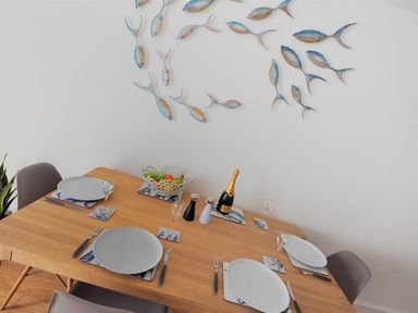 A dining table set for four with plates glasses cutlery a fruit bowl salt and pepper shakers a bottle of champagne and fish wall art at The Lower Deck in Lyme Regis