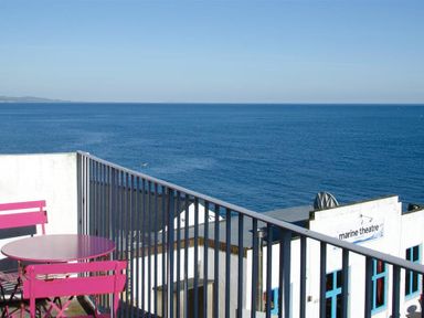 A balcony with pink table and chairs overlooking the ocean and a marine theatre building at 9 Bay View Court in Lyme Regis