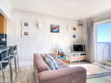 A living room with a pink sofa and pillows a small blue table a television and a balcony door showing the sea at 9 Bay View Court Lyme Regis