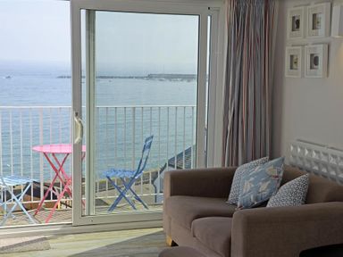 A living room with a brown sofa and cushions next to a sliding glass door opening to a balcony with a red table and blue chairs overlooking the sea at 9 Bay View Court in Lyme Regis