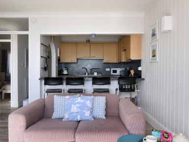 A view of a living room sofa with three cushions facing a kitchen with wooden cabinets and black bar stools at 9 Bay View Court in Lyme Regis