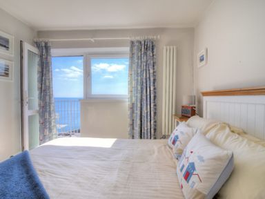 A bedroom with a bed with pillows and a blanket near a window with curtains overlooking the sea at 9 Bay View Court in Lyme Regis
