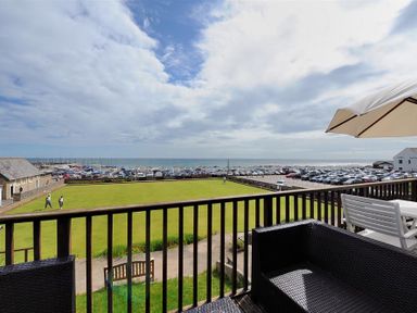 A balcony with outdoor seating and an umbrella overlooking a lawn with people and a parking area by the sea at 8 Bowling Green Chalets in Lyme Regis