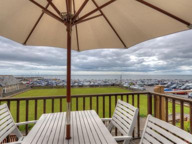 A balcony with a table umbrella and chairs overlooking a grassy area parking and the sea at 8 Bowling Green Chalets Lyme Regis