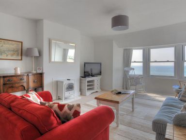 A living room with a red sofa, wooden coffee table, armchair by bay windows with ocean view, tv on cabinet and wooden sideboard at Ross House in Lyme Regis