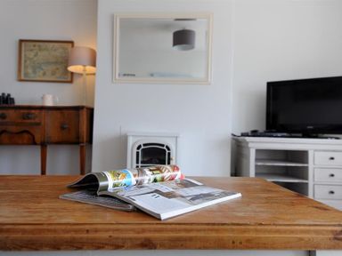 A wooden table with magazines in a living room with a lamp on a sideboard mirror above a fireplace and a television on a cabinet at Ross House in Lyme Regis