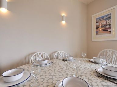 A dining table set with bowls, plates, and glasses with white chairs and a wall painting in the background at Ross House in Lyme Regis