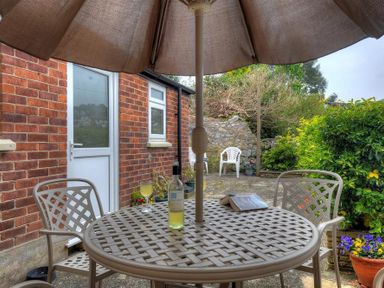 An outdoor patio with a round table under an umbrella surrounded by chairs and a brick wall with plants and flowers at 23 Lym Close Lyme Regis