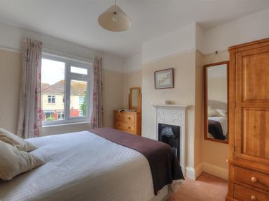 A bedroom with a bed a window with floral curtains a wooden wardrobe a chest of drawers a mirror and a fireplace at 23 Lym Close in Lyme Regis