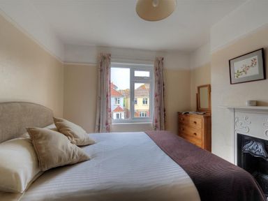 A bedroom with a bed a window with floral curtains a wooden dresser a framed picture and a fireplace at 23 Lym Close Lyme Regis