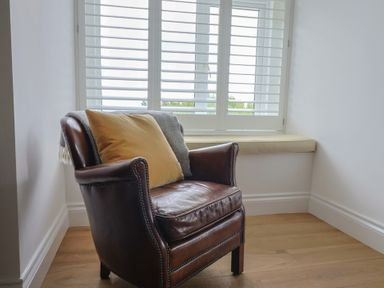A leather armchair with a beige cushion and a gray throw in front of a window with white shutters and a cushioned window seat in a room with wooden flooring at Ancient Lights in Lyme Regis