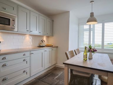 A kitchen with gray cabinets a wooden table with chairs a bottle and flowers near a window at Ancient Lights in Lyme Regis