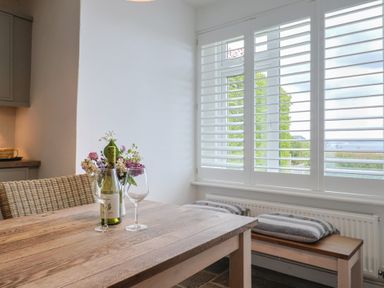 A wooden dining table with a bottle of wine two wine glasses and a vase of flowers next to wicker chairs and striped cushions on a bench by a window with white shutters at Ancient Lights in Lyme Regis