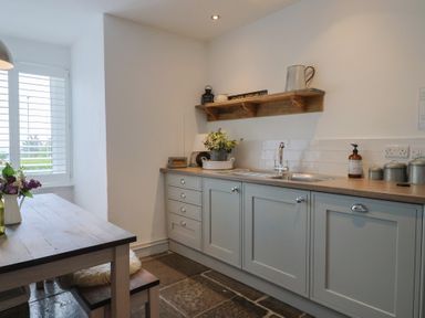 A kitchen with gray cabinets a wooden countertop a sink a wooden dining table with flowers and a window with white shutters at Ancient Lights in Lyme Regis